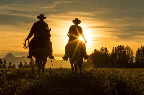 Two Cowboys Riding Into The Sunset Across Grassland With Mountains