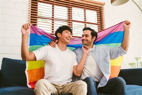Happy Caucasian And Asian Lgbt Couple Sitting On The Sofa Holding And