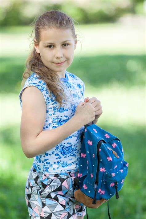 Happy Young Blonde Hair Girl Standing In Park With Small Backpack In Hands Looking At Camera