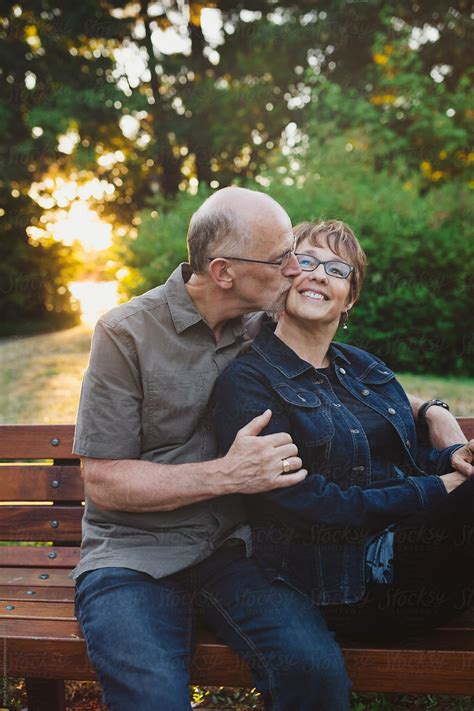 Middle Aged Retired Couple Kissing And Cuddling Outside Sitting On A Bench By Stocksy