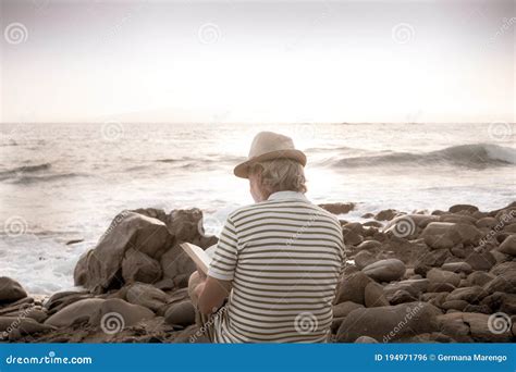 Senior Man With Hat Sitting On The Pebble Beach Reading A Book White Hair And Beard Stock Photo