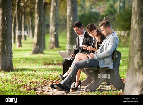 Three Business People Sitting On A Park Bench Using Laptop Smartphone