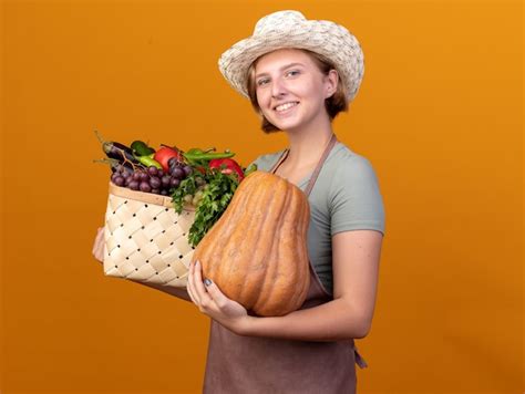 Молодой садовник в фартуке и шляпе держит ящик полный овощей и грозди винограда смотрит со