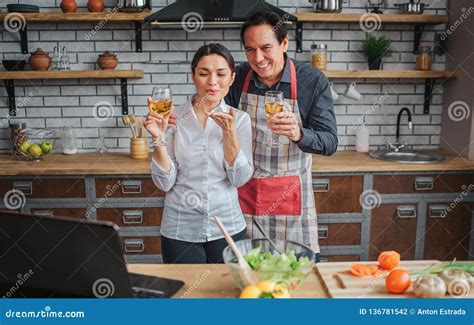 Cheerful Couple Stand Together In Kitchen They Hold Glasses Of Wine And Look On Laptop People
