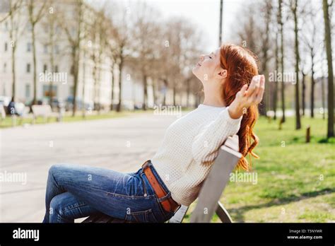 Young Woman In White Sweater And Blue Jeans Sitting On Bench In The