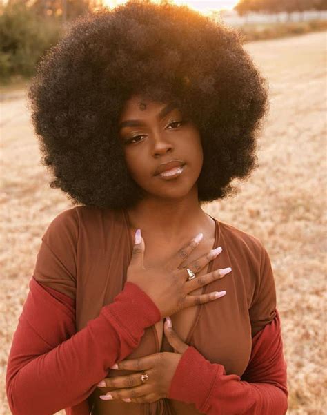 a woman with an afro standing in a field holding her hands on her chest and looking at the camera