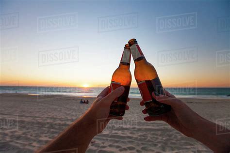 Close Up Of Hands Of Two People Doing Celebratory Toast With Beer Bottles On Beach Under Clear