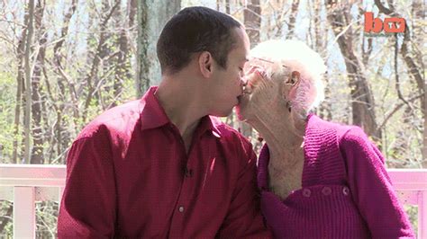 An Older Couple Kissing Each Other While Sitting On A Bench In Front Of Some Trees