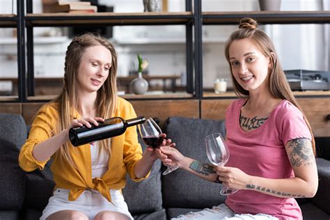 two lesbians holding wine glasses and pouring wine while sitting on sofa in living room stock