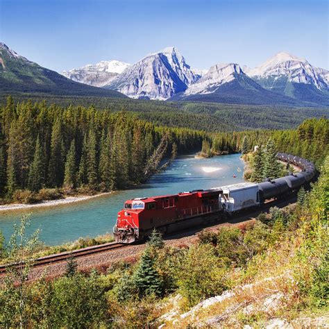 A Train Traveling Down Tracks Next To A River And Mountain Range With