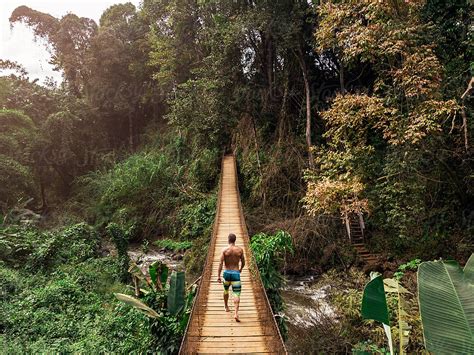 Young Man Walking On Footbridge Above River Between Tropical Forest