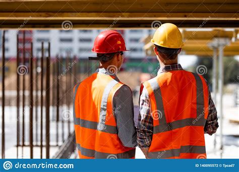 Two Civil Engineers Dressed In Orange Work Vests And Helmets Discuss