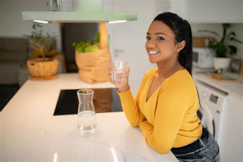 Happy Young African American Woman Sit At Table Drinking Water From Glass In Modern Kitchen