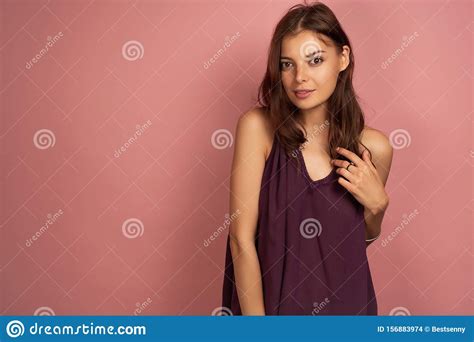 A Brunette Stands On A Pink Background In A Purple Top Looking At The