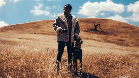 Travis Scott Is Standing With Dog On Dry Grass In Cloudy Sky Background