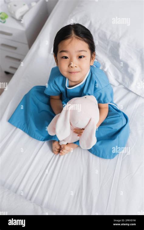 Top View Of Little Asian Girl Sitting On Hospital Bed With Toy Bunny