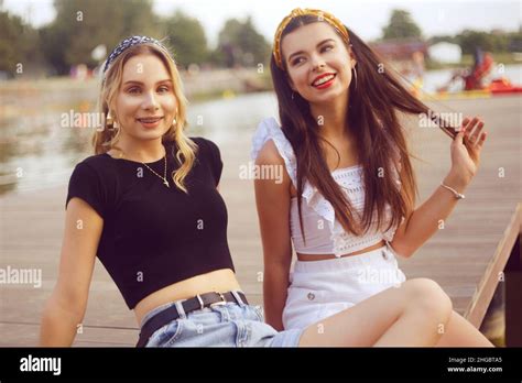 Two Fashionable Girls Girlfriends Are Sitting On The Pier Near The River The Beach Girls