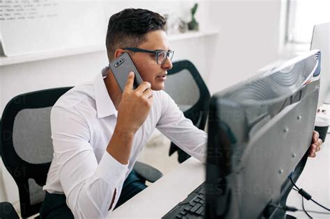 Software Developer Talking Over Mobile Phone Sitting At His Office Desk With A Desktop Computer