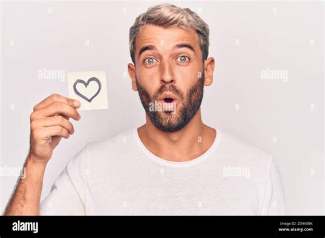 Handsome Blond Man With Beard Holding Reminder With Heart Shape Over White Background Scared And