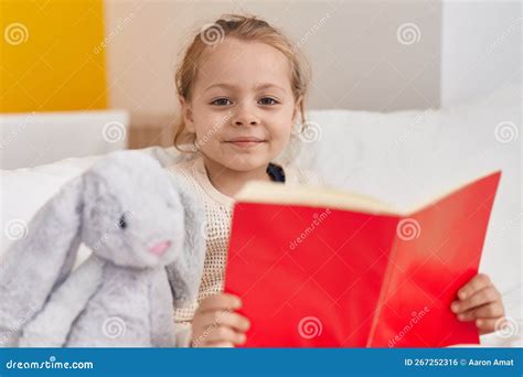Adorable Blonde Girl Reading Book Sitting On Bed At Bedroom Stock Photo
