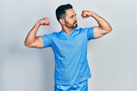 Handsome Hispanic Man With Beard Wearing Blue Male Nurse Uniform Showing Arms Muscles Smiling