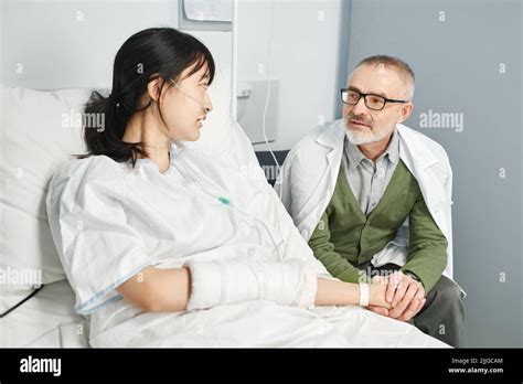 Loving Father Visiting His Daughter In Hospital Sitting On Chair Holding Her Hand And Chatting