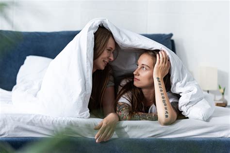 Two Lesbians Lying Under Blanket And Looking At Each Other In Bedroom