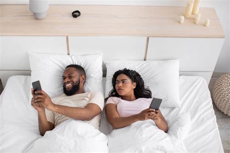 disgruntled millennial african american wife looks at husband smartphone on bed in bedroom