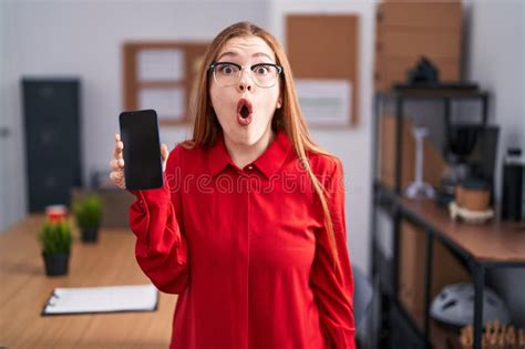 Redhead Woman Working At The Office Showing Smartphone Screen Scared