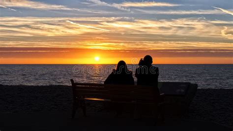 A Romantic Scene A Couple In Silhouette Sit On A Bench By The Sea At