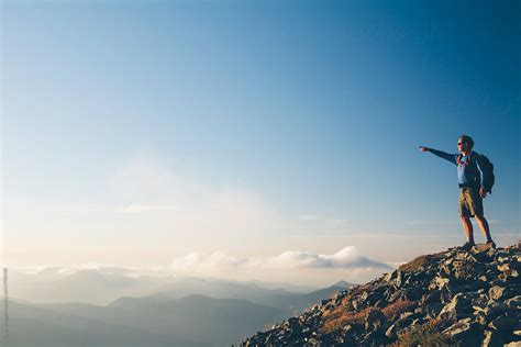 Man Standing On Mountain Summit Pointing Towards Mountains And Sky
