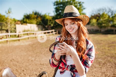 Smiling Happy Redhead Cowgirl Using Mobile Phone While Standing At Ranch Fence Royalty Free