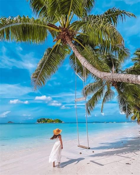 A Woman In A White Dress And Straw Hat Walks On The Beach Near Palm Trees