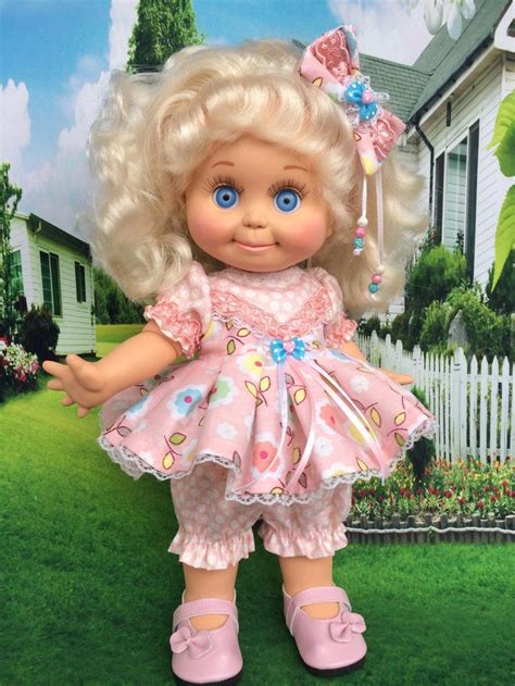a doll with blonde hair and blue eyes standing in front of a white picket fence