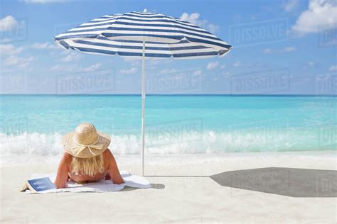 Woman Relaxing On Sunny Beach With Book Under Striped Beach Umbrella
