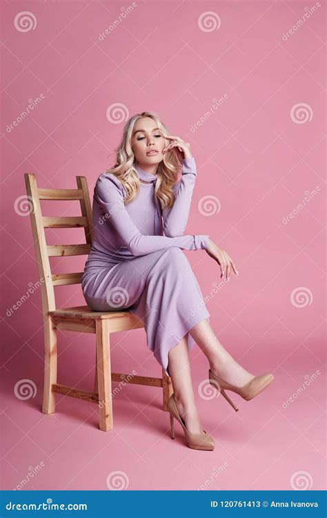 Fashion Beautiful Blonde Woman In Knitted Closed Long Dress Sitting On A Chair On A Pink