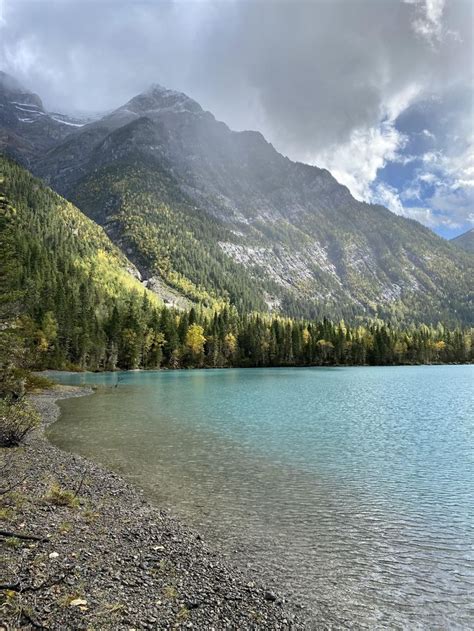 A Lake Surrounded By Mountains And Trees Under A Cloudy Sky