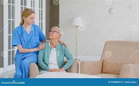 Smiling Blonde Nurse In Uniform Takes Care Of Senior Woman In Light Room At Assisted Living