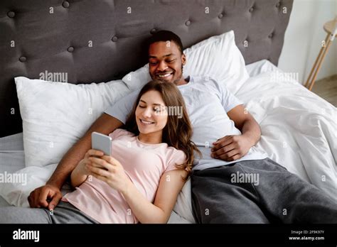 happy interracial couple lying on bed with smartphone browsing web having online video call