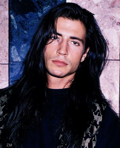 A Man With Long Black Hair Standing In Front Of A Tile Wall And Looking