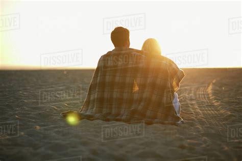 couple on beach wrapped in blanket watching sunset rear view stock
