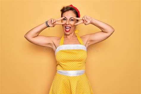 middle age senior pin up woman wearing 50s style retro dress over yellow background doing peace
