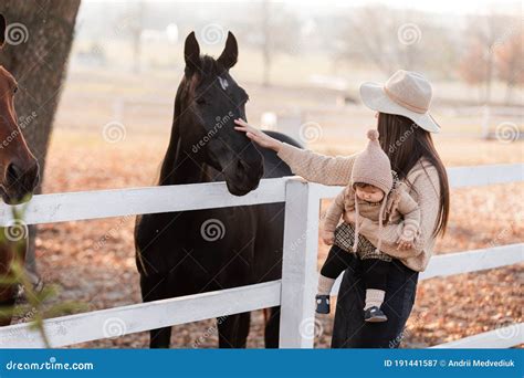 Young Mother And Little Baby Girl Near A Horses In Autumn Sunny Day
