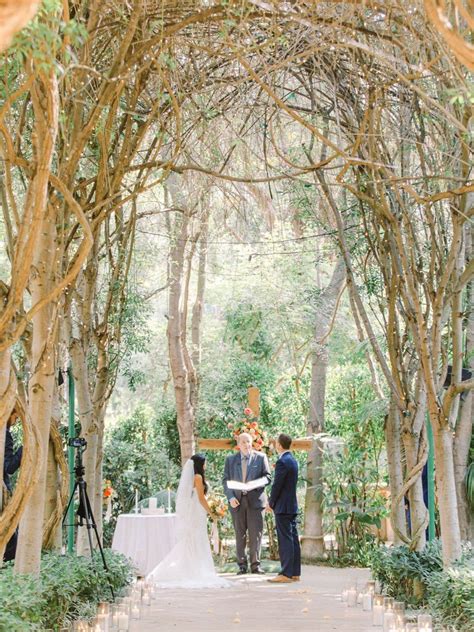 A Bride And Groom Are Standing Under An Arch Made Of Trees With Candles
