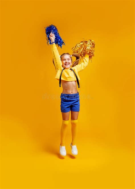 cute cheerleader girl in a yellow tank and blue shorts holds pompons and dances jumps stock