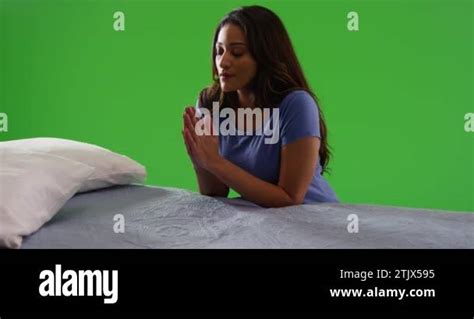 Young Latina Woman Kneeling By Her Bed Praying With Hands Pressed Together On Green Screen On