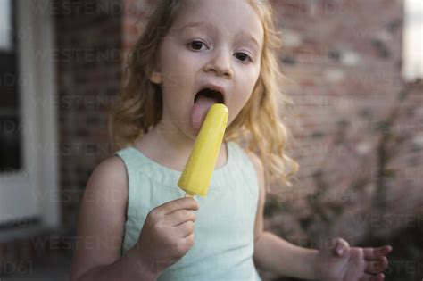 Portrait Of Girl Eating Popsicle Stick While Standing Against House Stock Photo