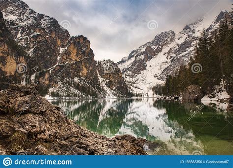 Rocks On The Water At Braies Lake And Dolomite With Snow In A Cloudy