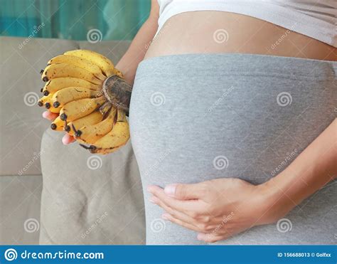 Pregnant Woman Stand In Room And Holding Banana At Her Belly Stock