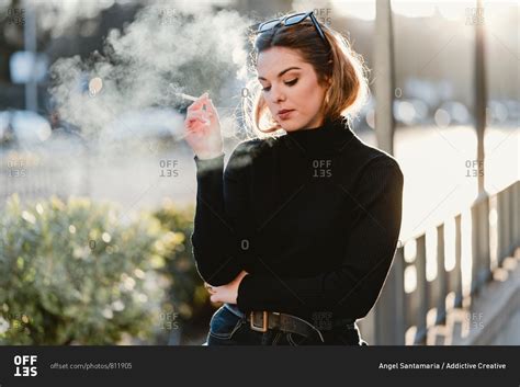 beautiful lady in trendy outfit exhaling fume while smoking cigarette on sunny day on city